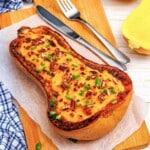 A roasted half butternut squash topped with pecans and chives on a wooden cutting board with a fork and knife.