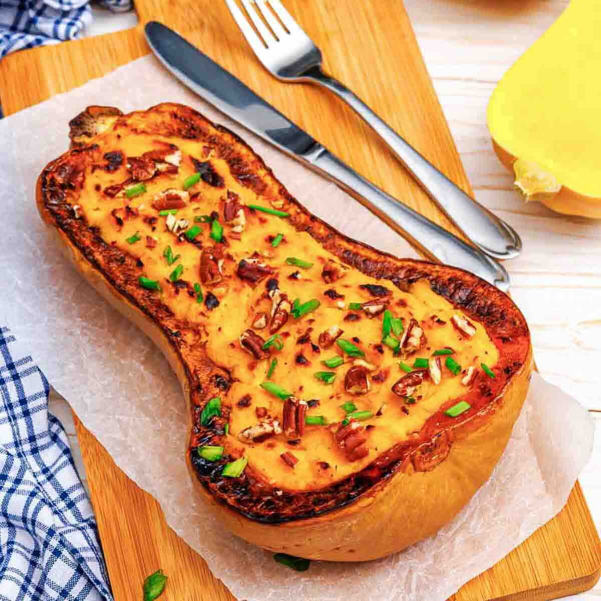 A roasted half butternut squash topped with pecans and chives on a wooden cutting board with a fork and knife.