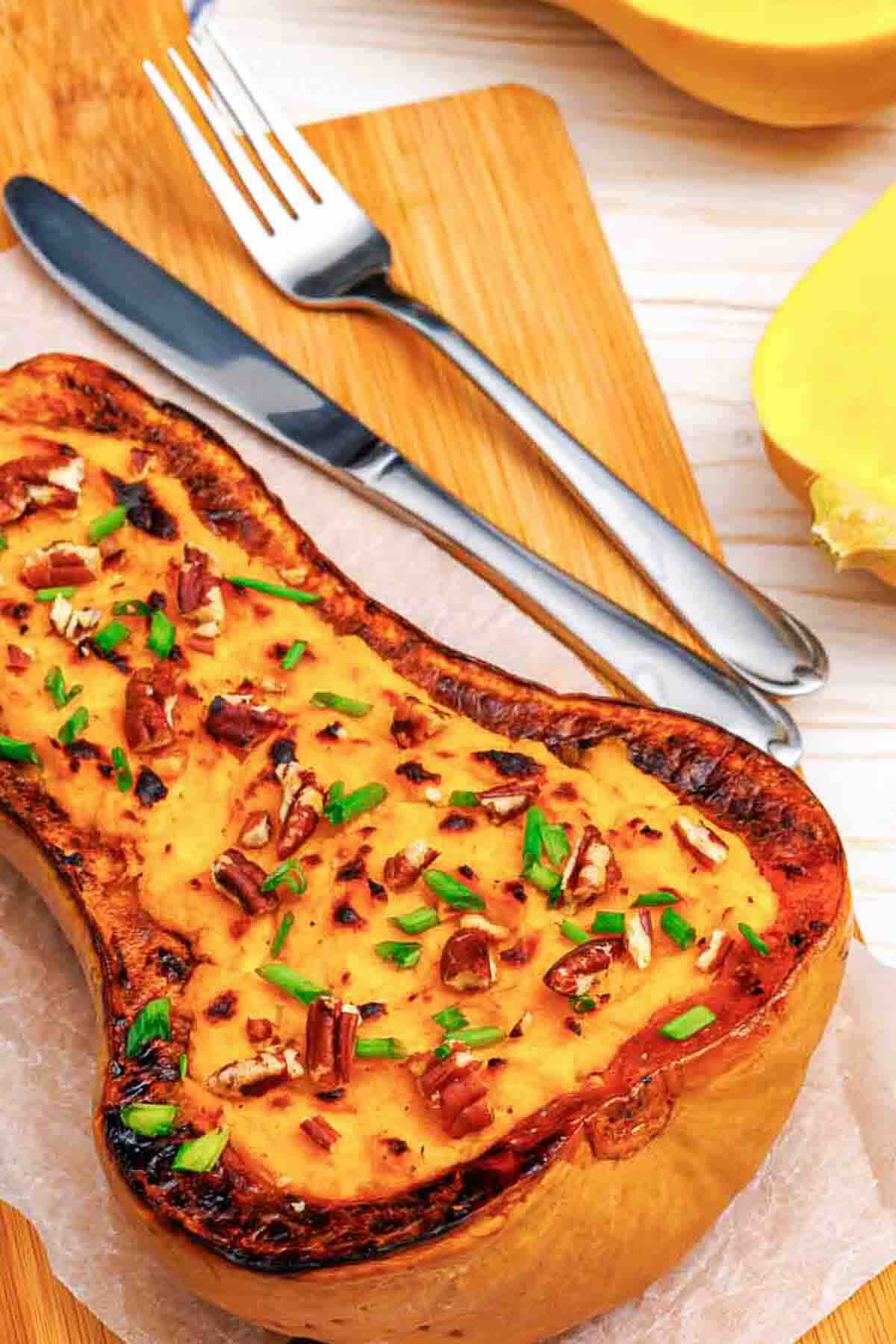 Roasted butternut squash topped with pecans and chives, served on a wooden cutting board with a fork and knife.