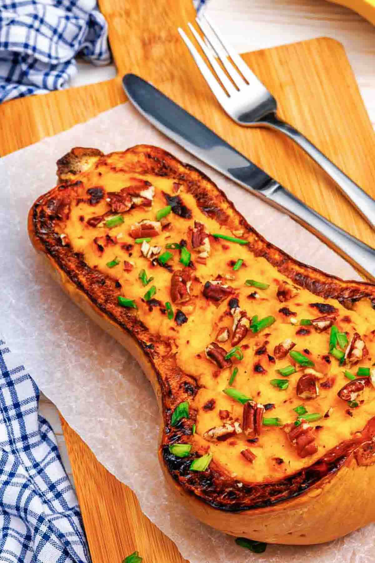 Roasted butternut squash half topped with pecans and chopped chives on a wooden board with a knife and fork.