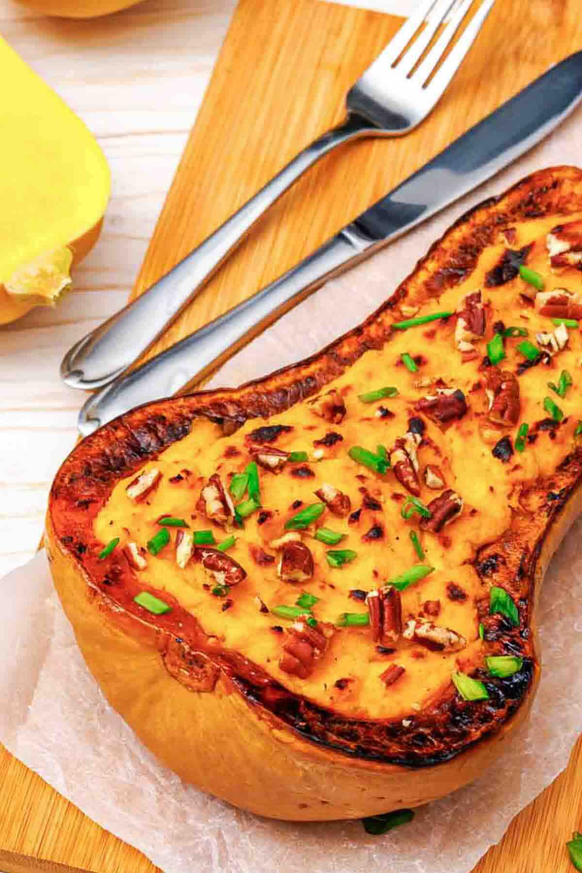 Roasted butternut squash stuffed with mashed squash, topped with pecans and chives, served on a wooden board with a fork and knife.