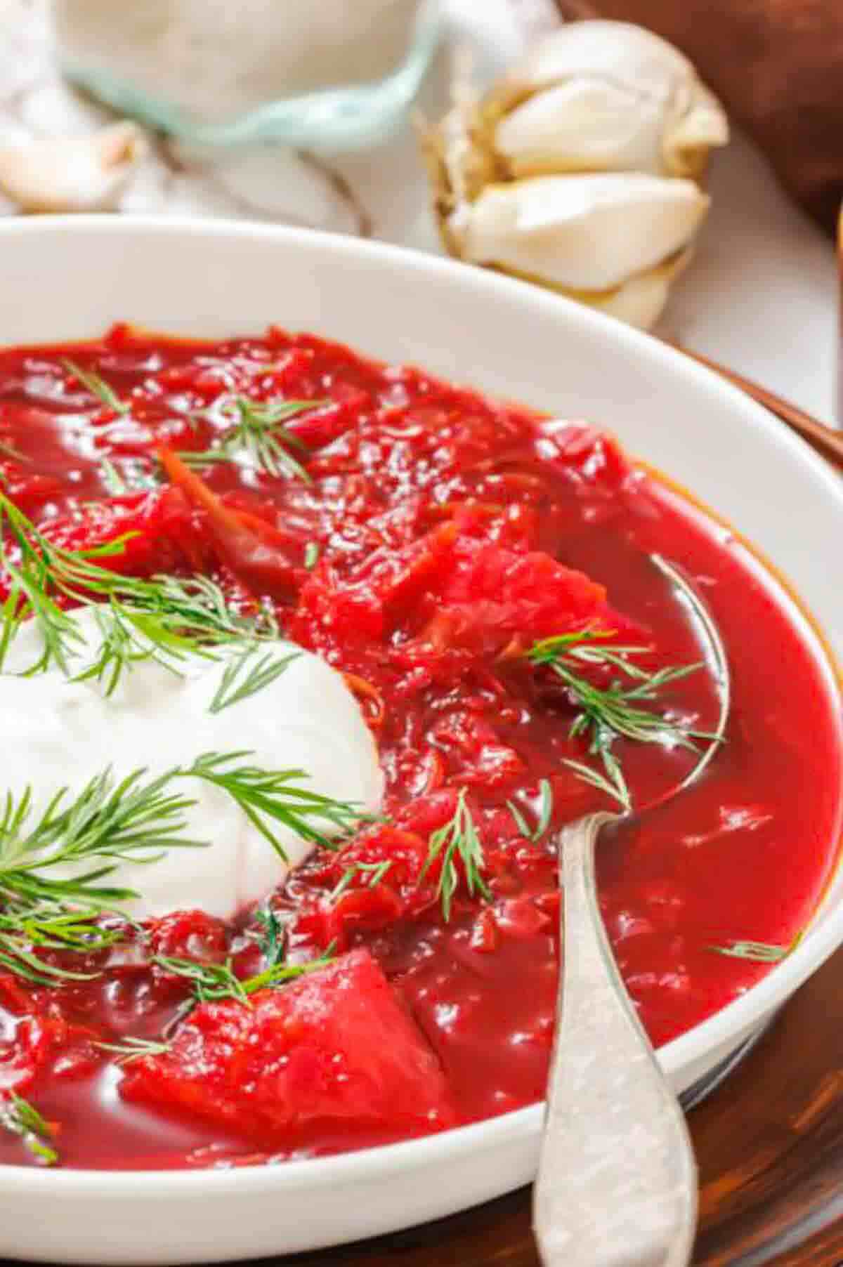 A bowl of red beet soup garnished with a dollop of sour cream and fresh dill, served with a spoon.
